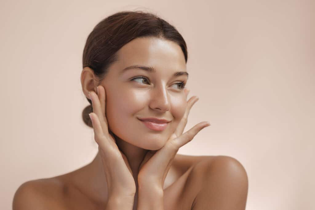 A woman with healthy, dewy skin looks off to the side, using her hands to frame the well-balanced features of her face after a liquid facelift.