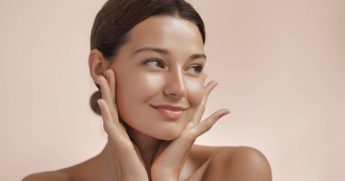 A woman with healthy, dewy skin looks off to the side, using her hands to frame the well-balanced features of her face after a liquid facelift.