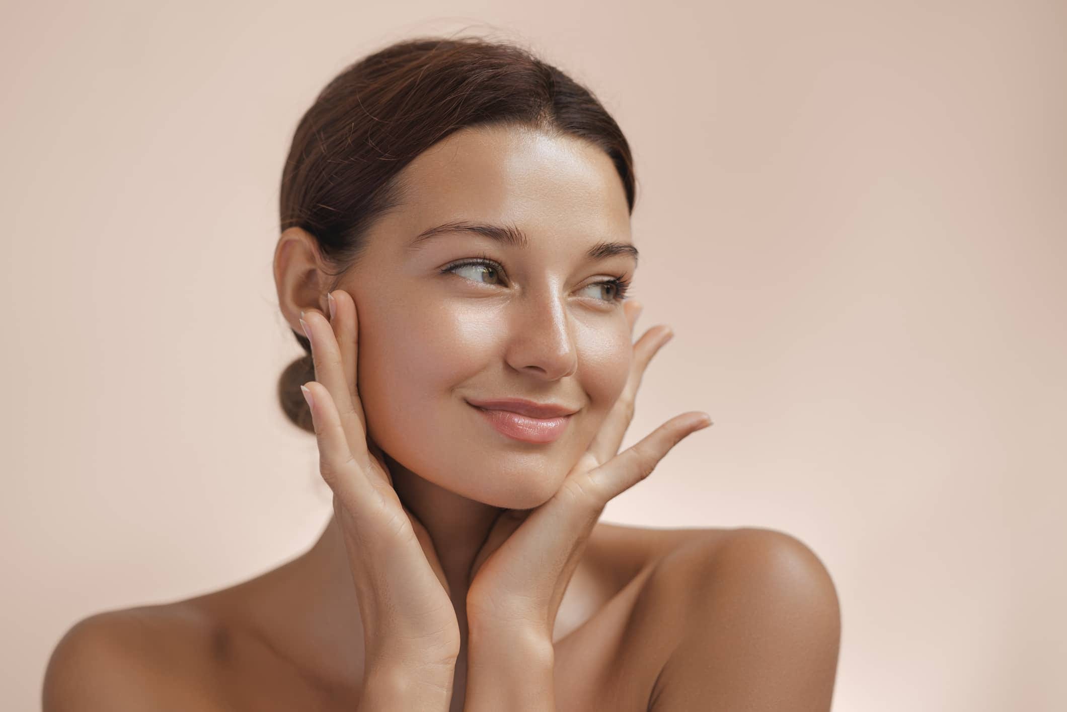 A woman with healthy, dewy skin looks off to the side, using her hands to frame the well-balanced features of her face after a liquid facelift.