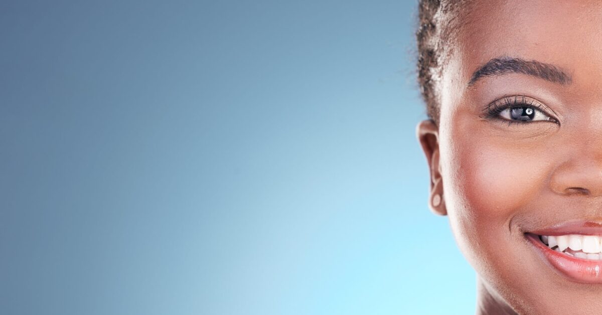 Partial view of a dark-skinned woman’s face. She smiles at the camera, proud of her beautiful, rejuvenated complexion.