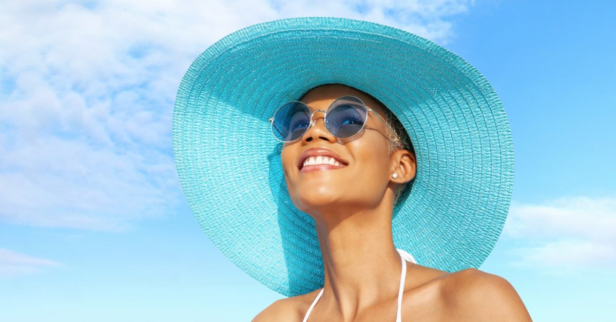 A woman wearing a straw hat and sunglasses stands in front of a blue sky, ready to enjoy the nice weather as she thinks about summer wellness tips.