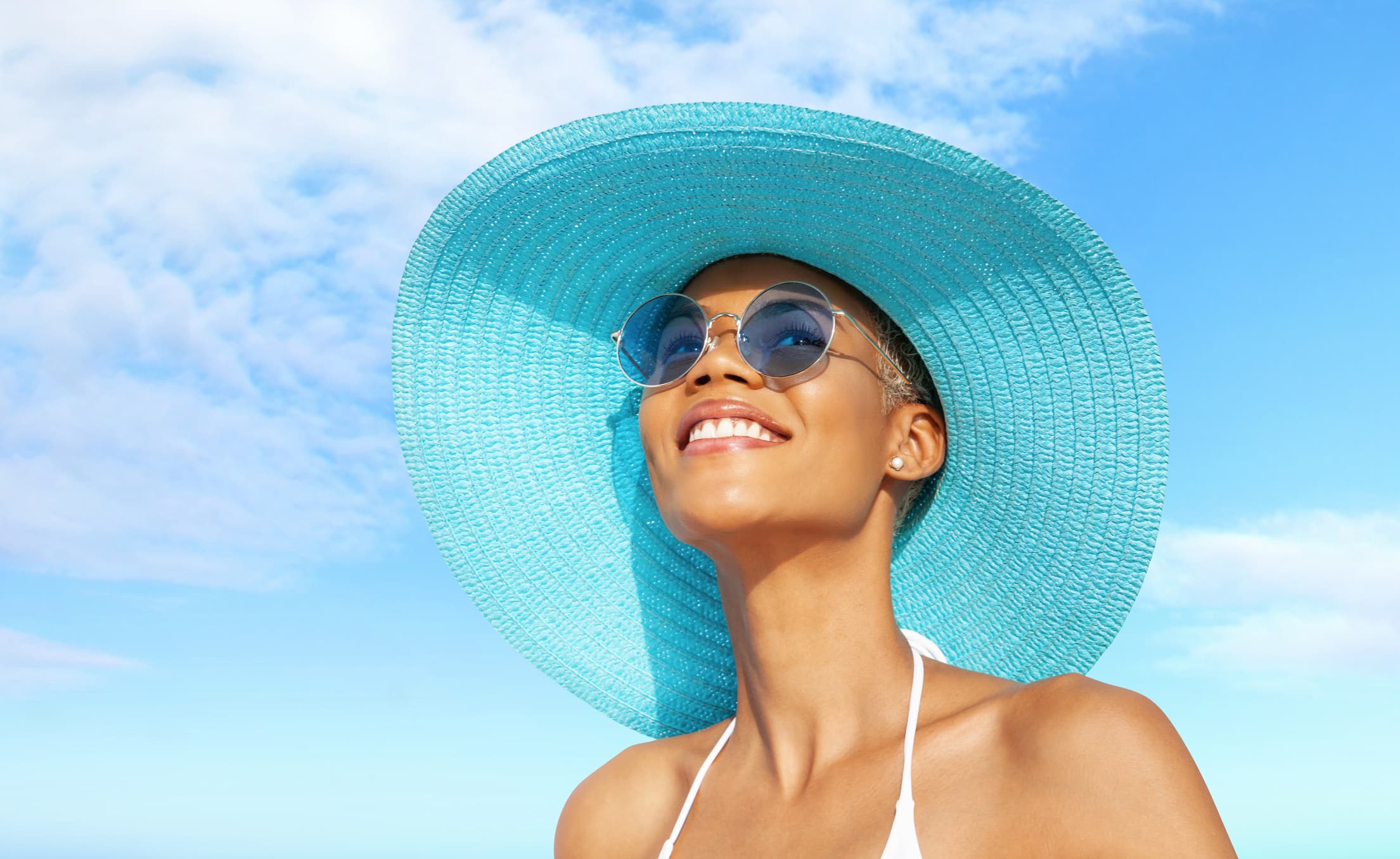 A woman wearing a straw hat and sunglasses stands in front of a blue sky, ready to enjoy the nice weather as she thinks about summer wellness tips.