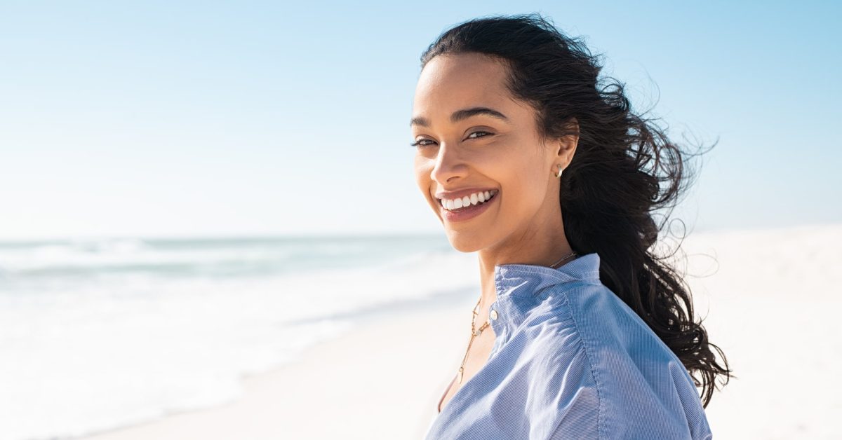 A young woman on a beach smiles at the camera, glad to enjoy her wrinkle-free vacation after tox injections.