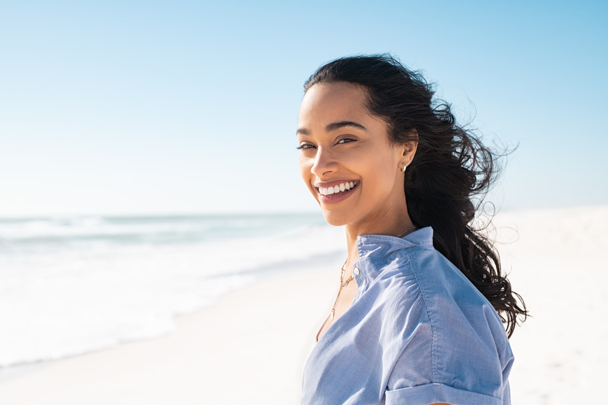 A young woman on a beach smiles at the camera, glad to enjoy her wrinkle-free vacation after tox injections.