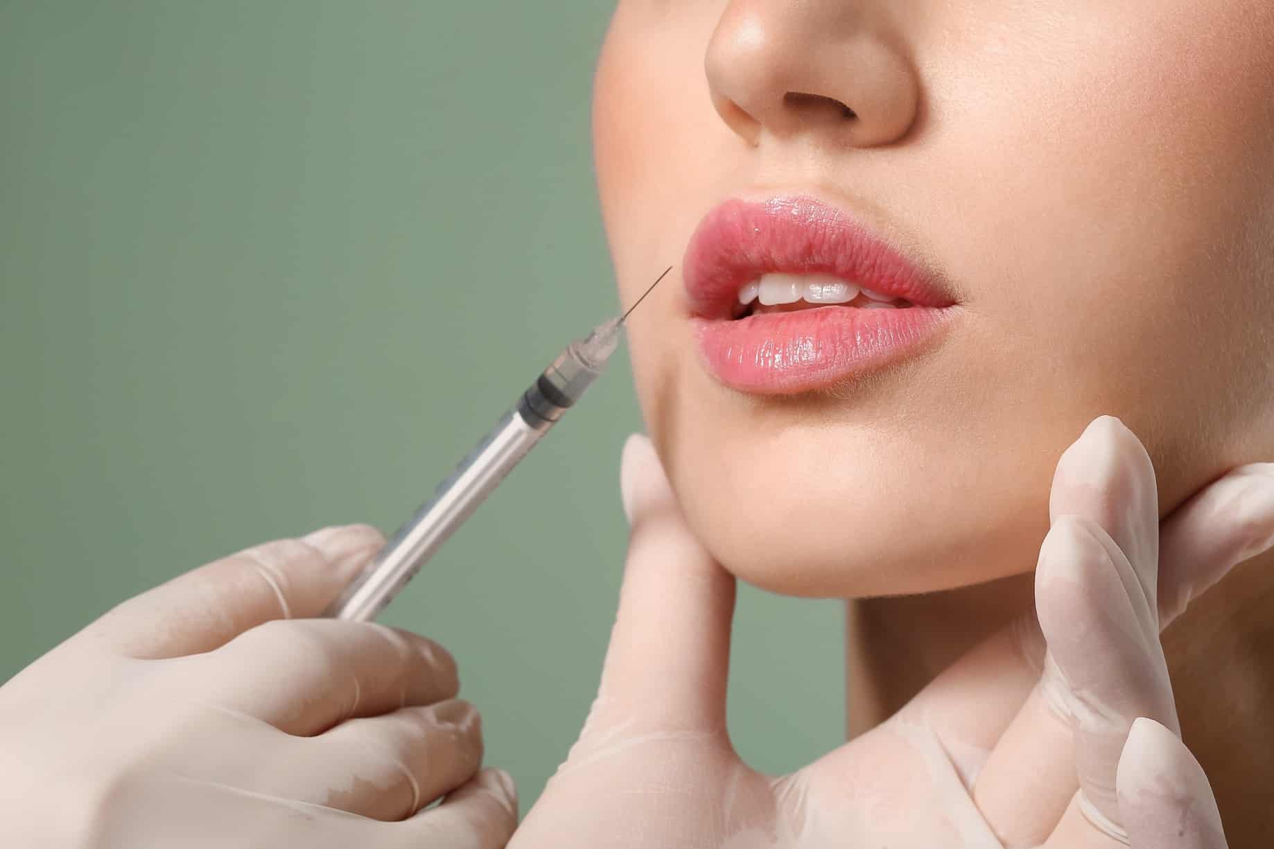 Close up of a woman’s face from the bottom of her nose to her chin and a gloved hand holding her chin while another prepares to inject her upper lip with a syringe.