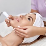 A woman wearing a white headband receives a facial skincare treatment from an esthetician using a specialized wand tool