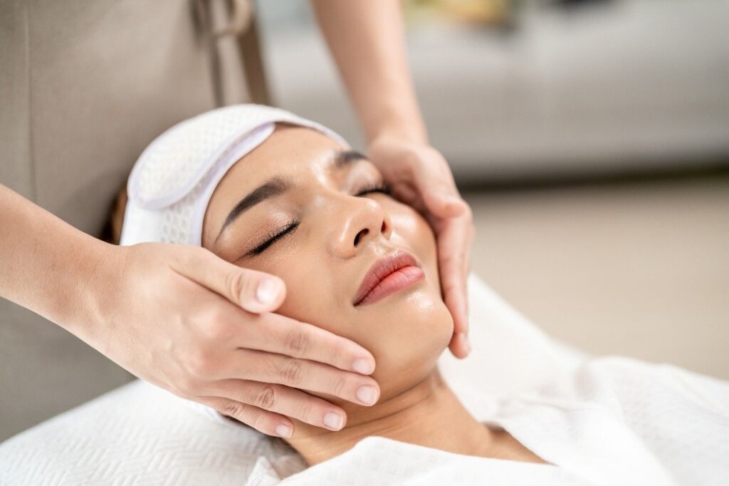 A woman with eyes closed and a white headband receives a relaxing facial massage from a spa professional.
